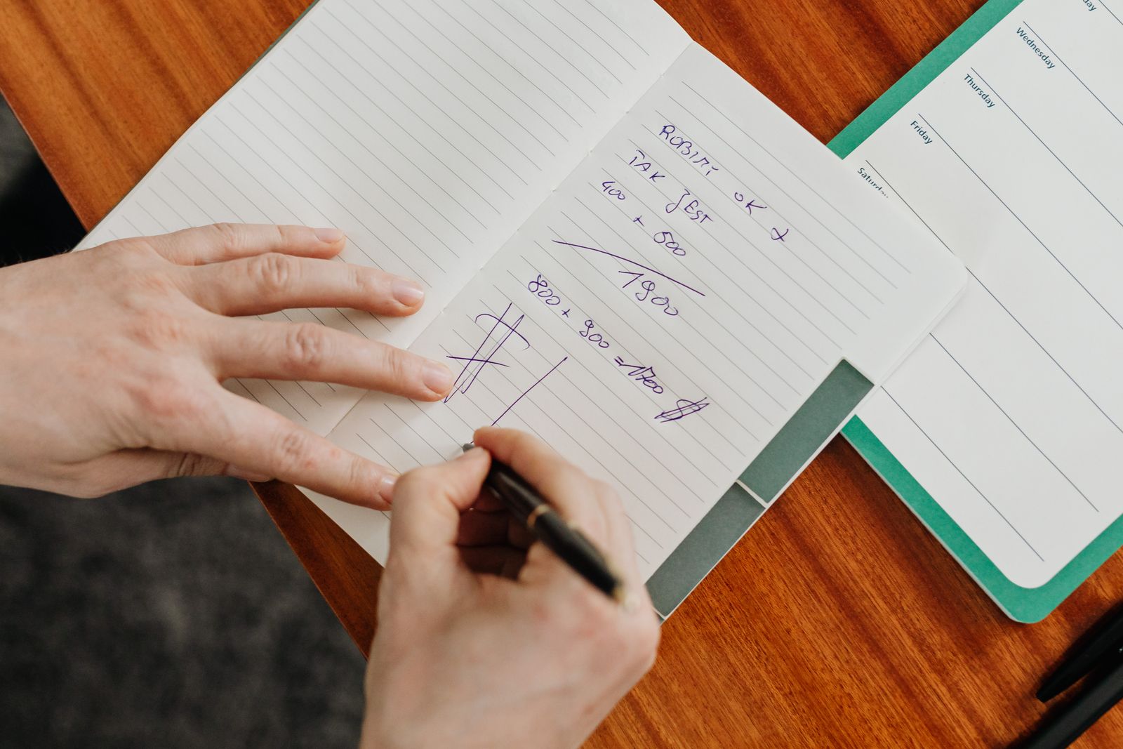 A hand writing sums in a notebook on a wooden desk