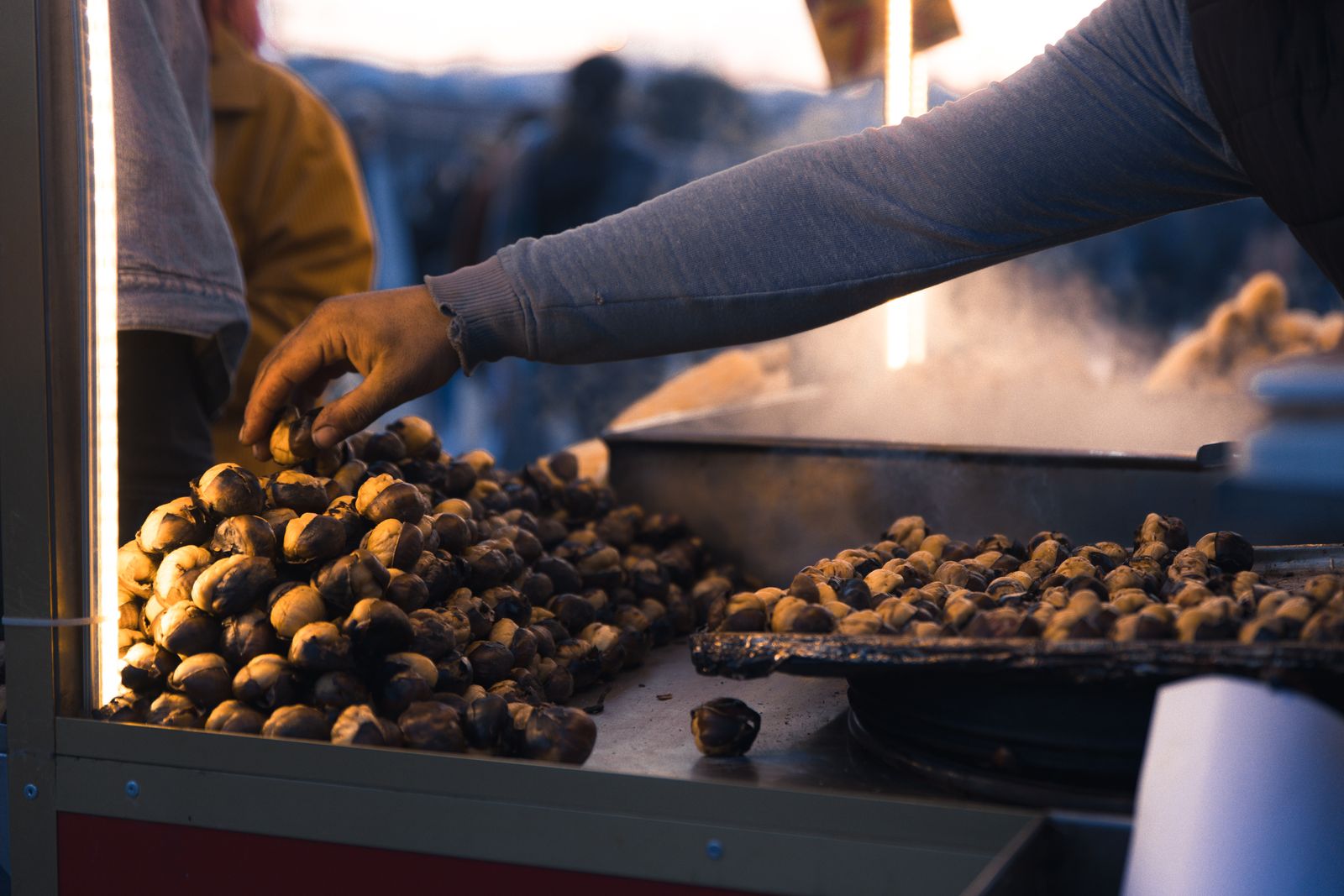 A stallholder's hand reaching over a pile of roasted chestnuts on the grill at night