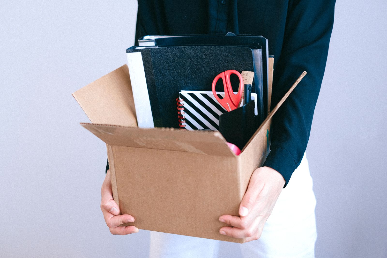 A woman carrying a cardboard box of desk things, her face out of frame