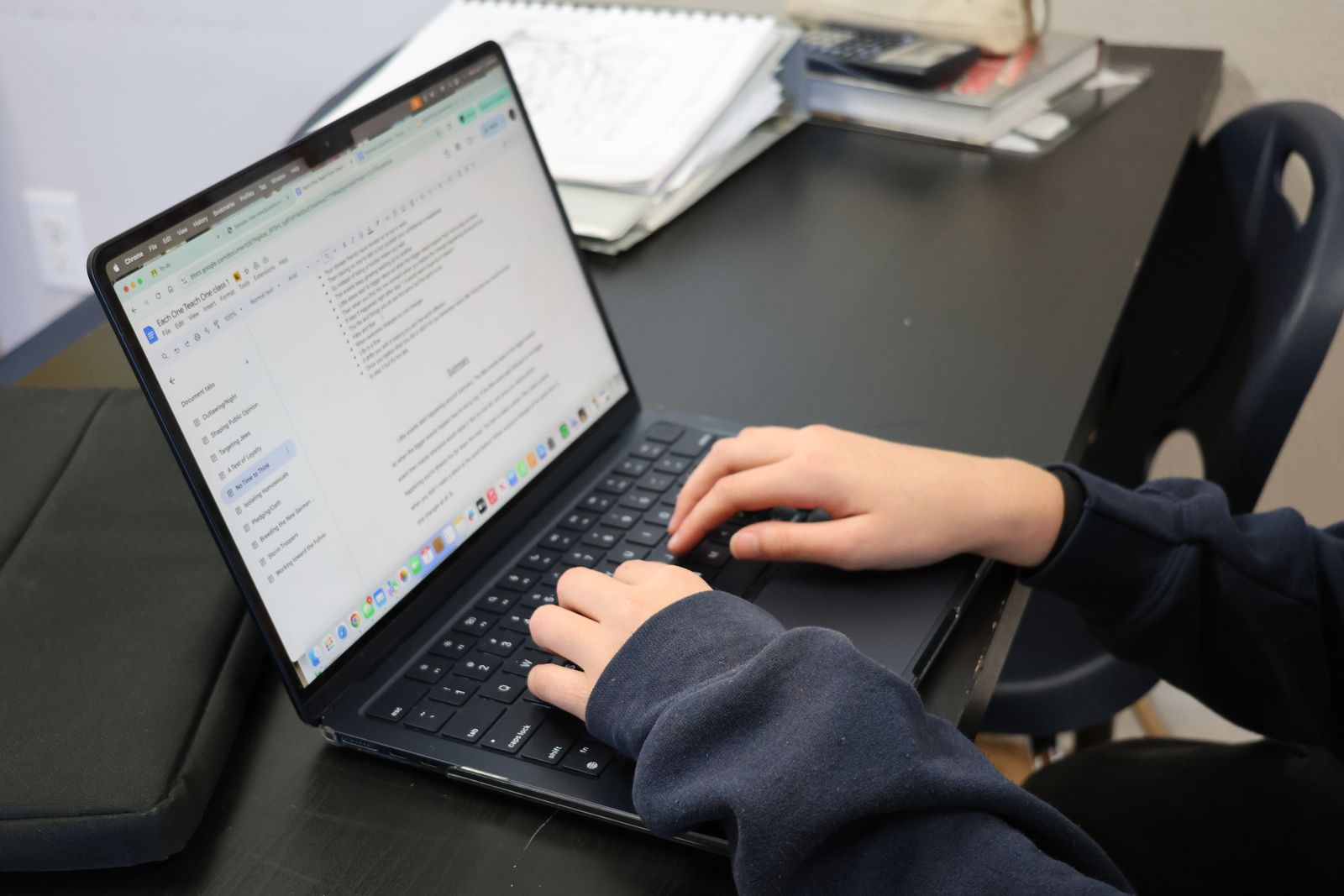 A person typing on a laptop at a desk with papers and books