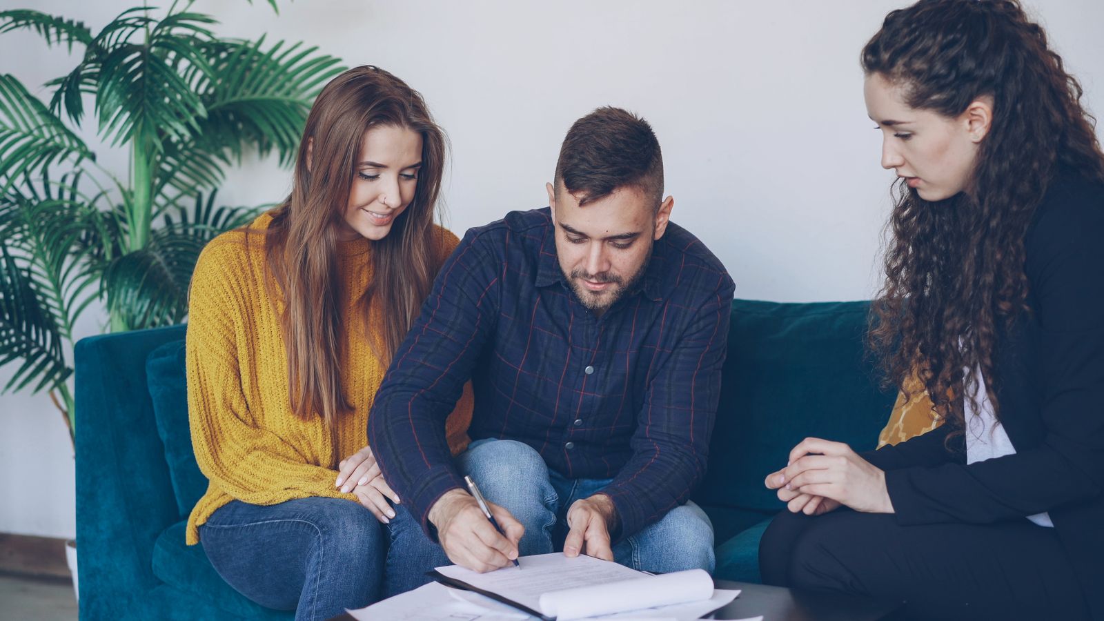 A young couple signing mortgage papers with an adviser on a sofa