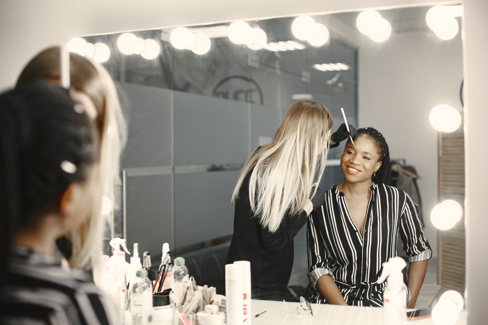 An artist does a client's makeup in front of a mirror with bulb lights