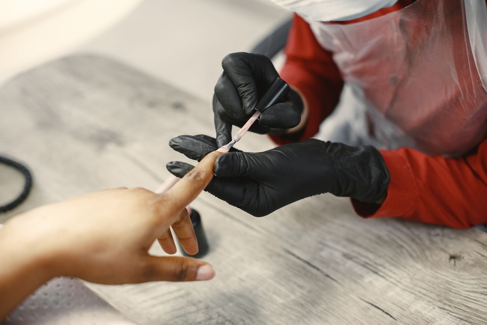 A nail technician in black gloves painting a client's nails at the desk