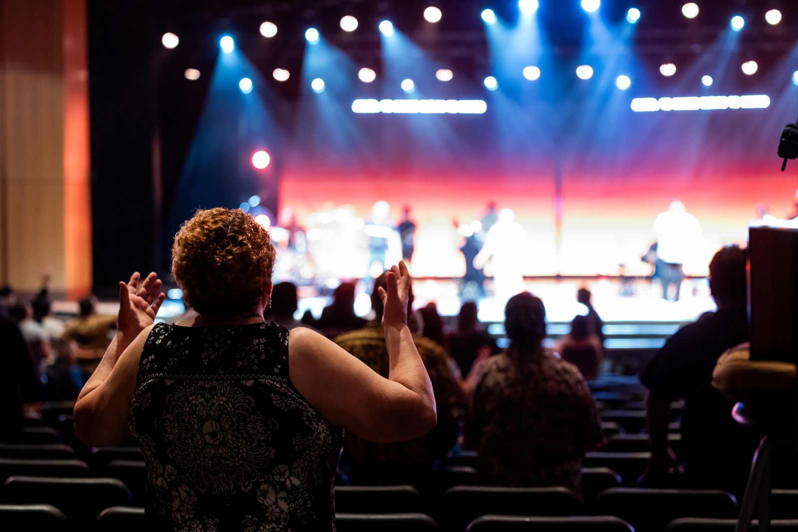 An audience clapping in front of a lit stage, seen from behind