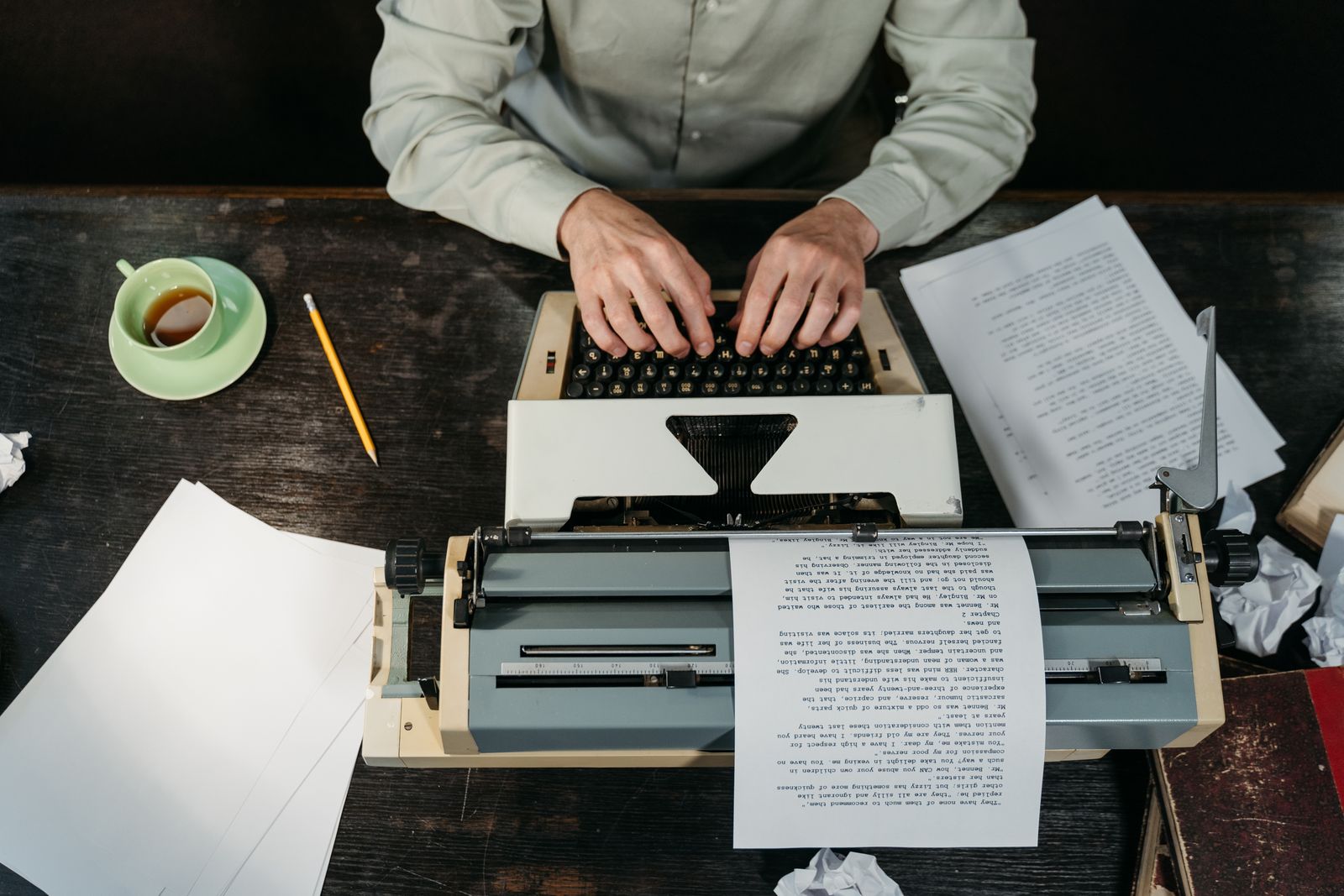 Hands typing on an old typewriter on a dark desk, with loose pages and a coffee cup