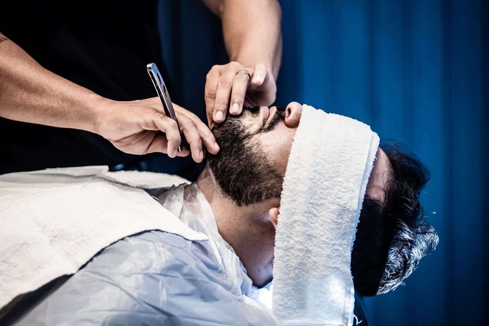 A client reclined under a towel while the barber shaves his beard line with an open razor