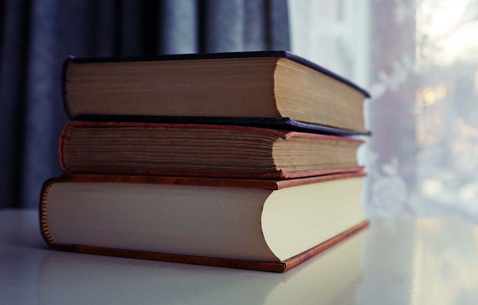 A stack of old hardback books on a shelf by a sunlit window