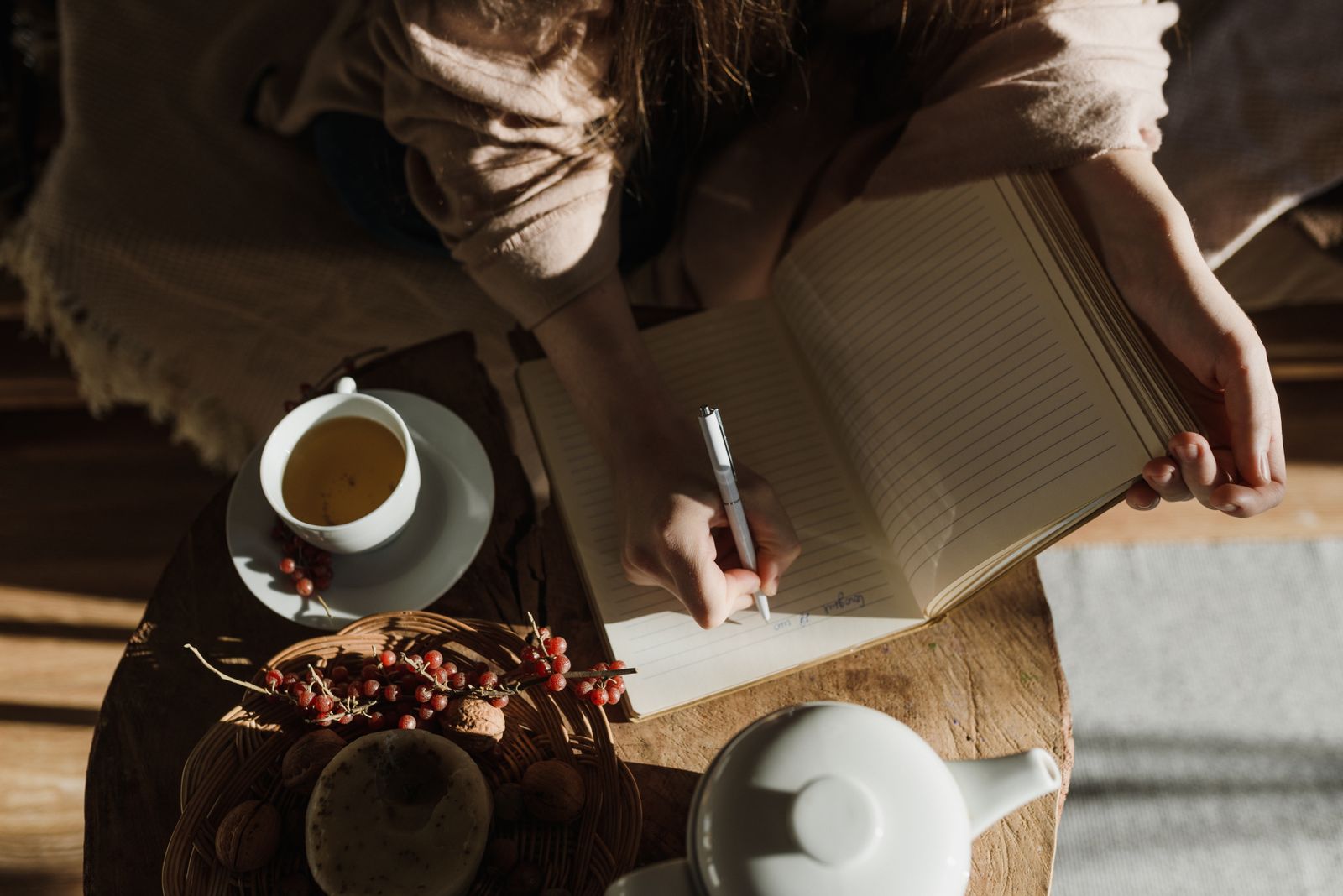 Hands writing in a notebook beside a cup of tea and a teapot