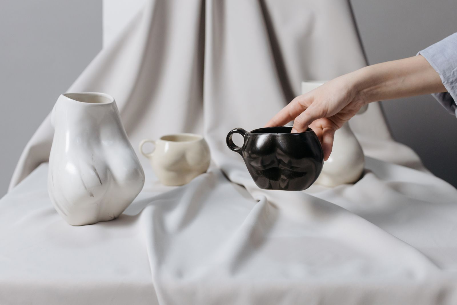 A hand placing a black ceramic teapot beside white vases on draped linen