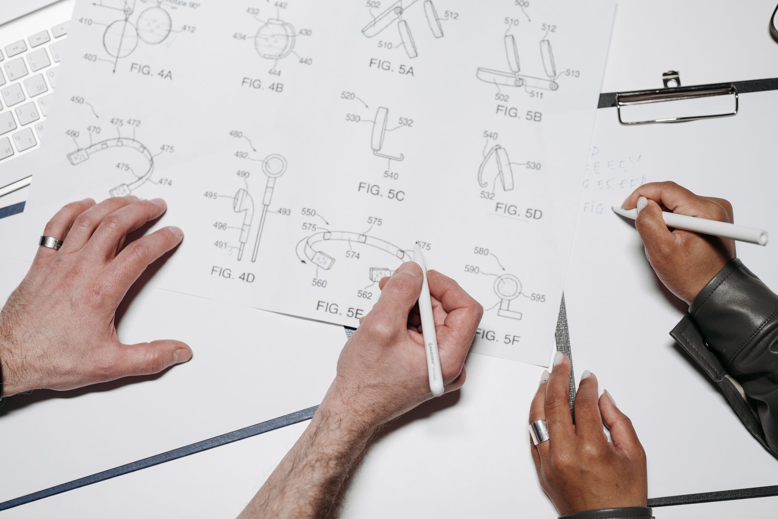 Two people's hands with pens over technical drawings on a white table