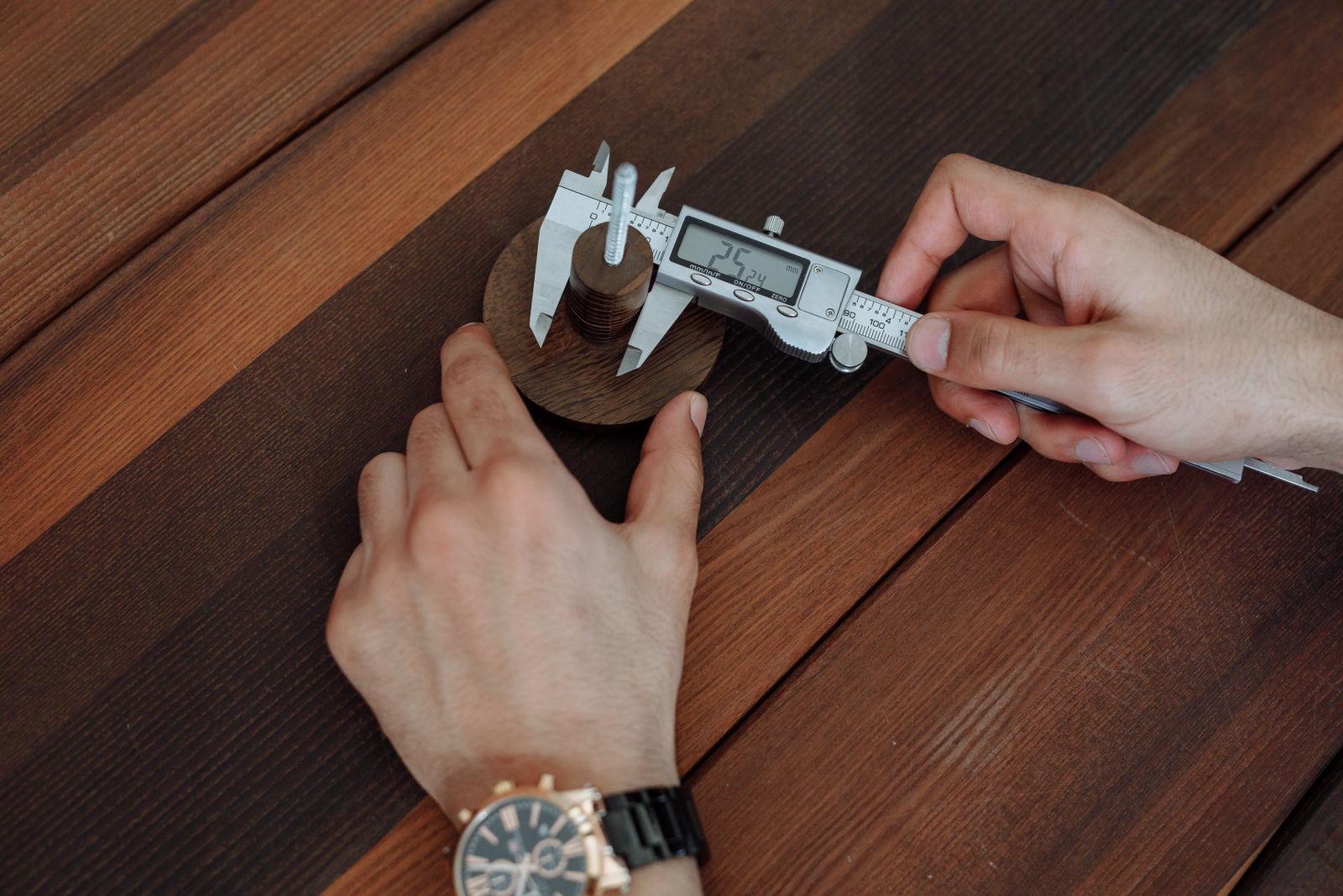 Hands measuring a small part with a digital caliper on a wooden table