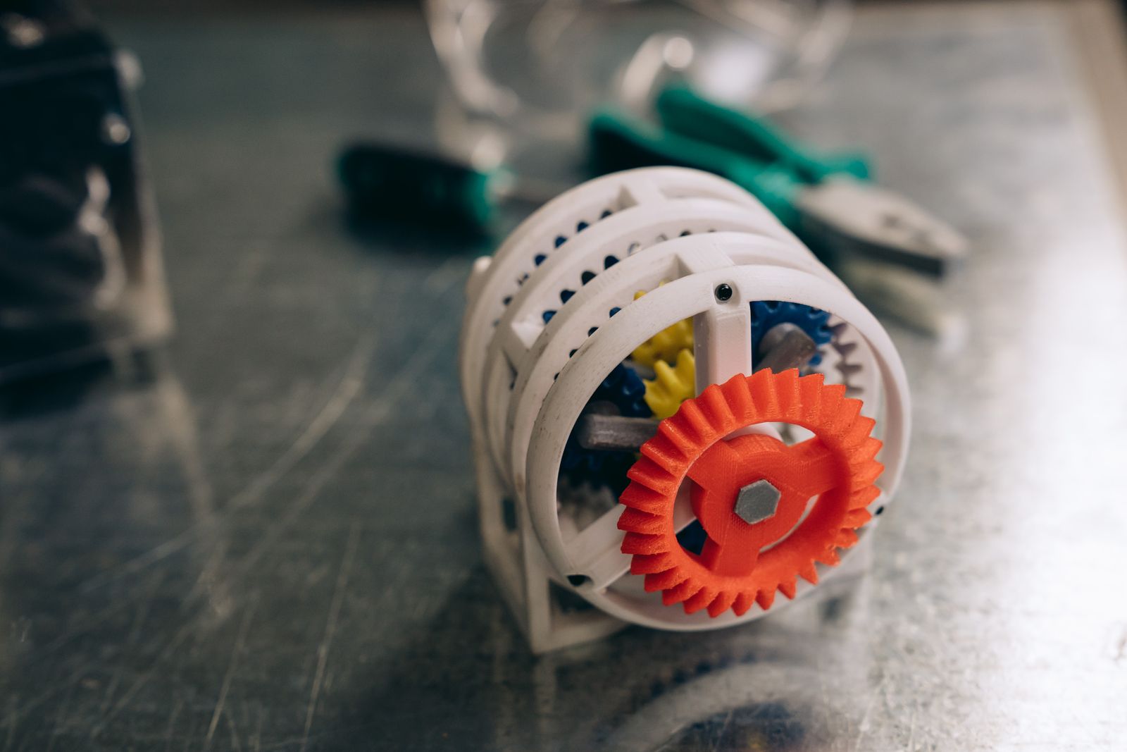 A 3D printed gear mechanism in white and orange on a metal bench