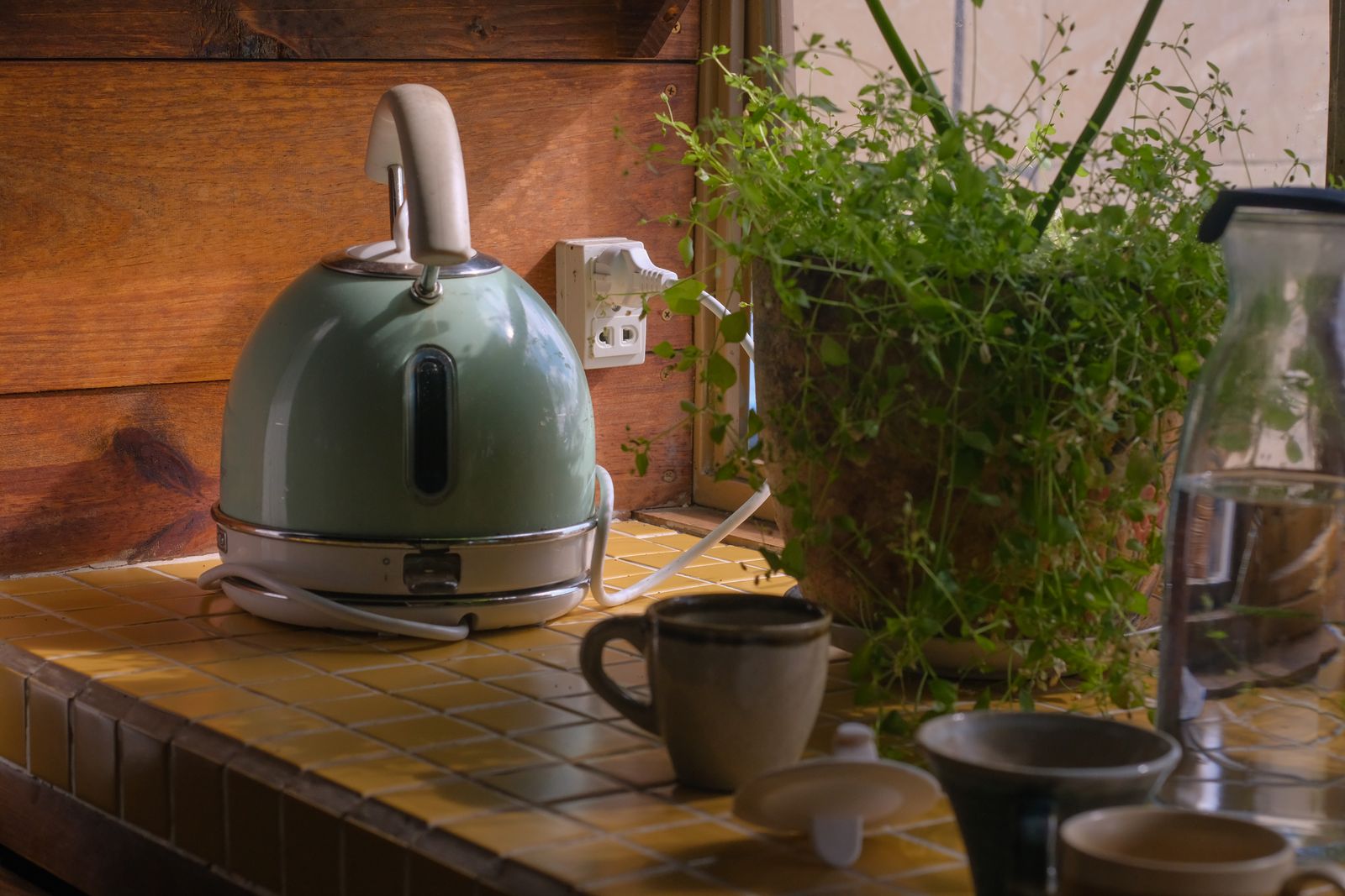 A pale green electric kettle on a tiled kitchen counter beside mugs and a potted herb