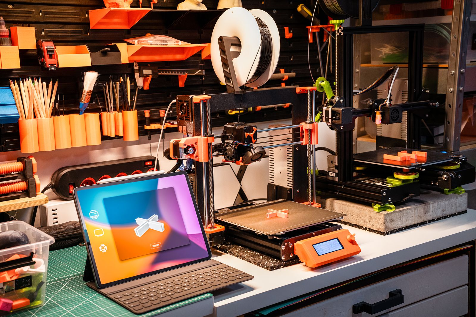A workbench with two 3D printers, a tablet on a green cutting mat and tools hung on the wall