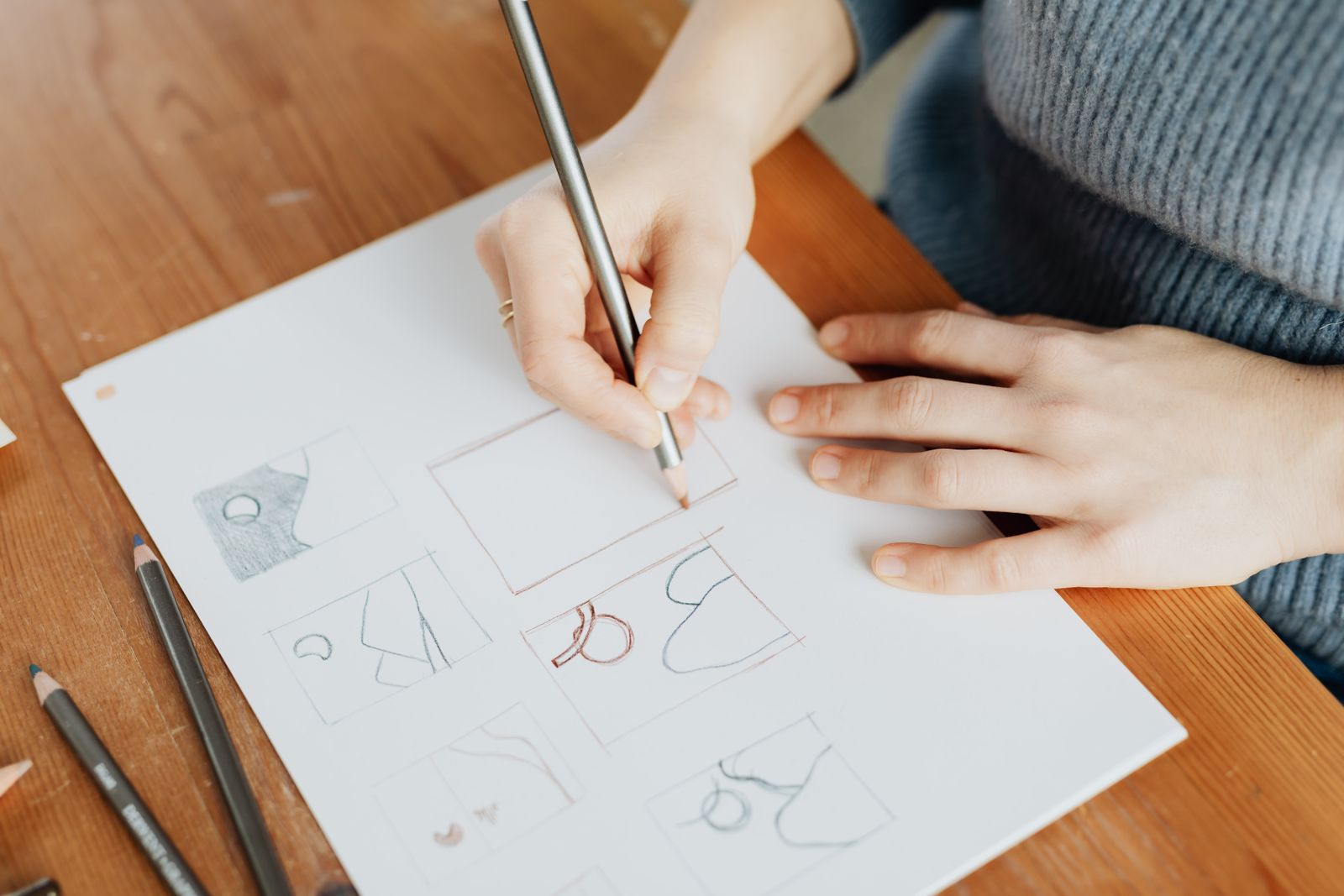 Seen from above, a designer in a knitted jumper sketches with a pencil at a wooden table