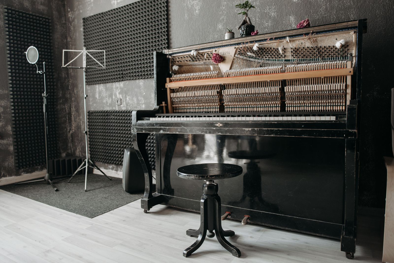 An open upright piano and its stool in a studio room lined with grey acoustic foam, a microphone stand and a music stand beside it