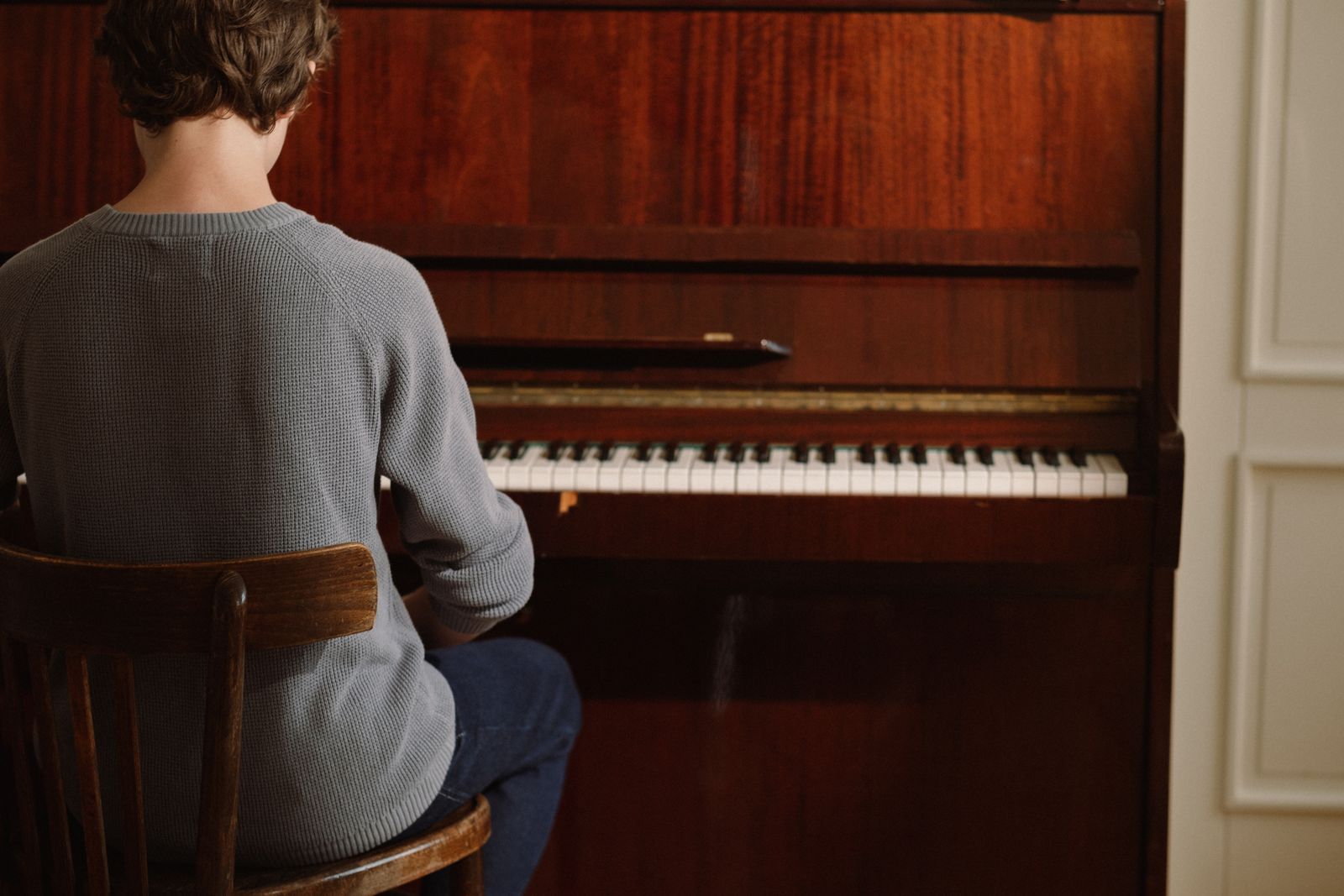 A pianist in a grey jumper seen from behind at a wooden upright piano