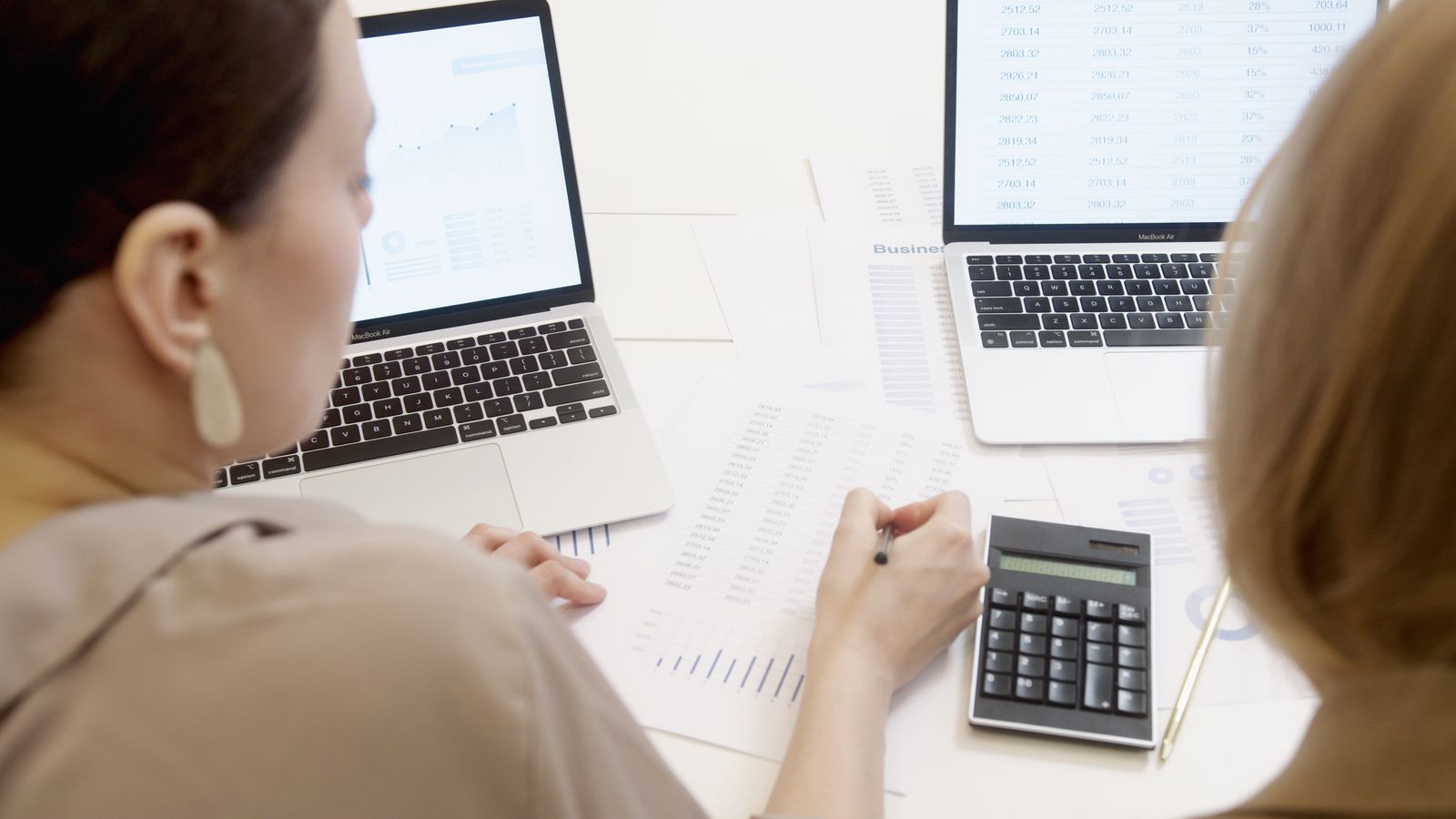 Two people at a desk with laptops, a calculator and printed figures