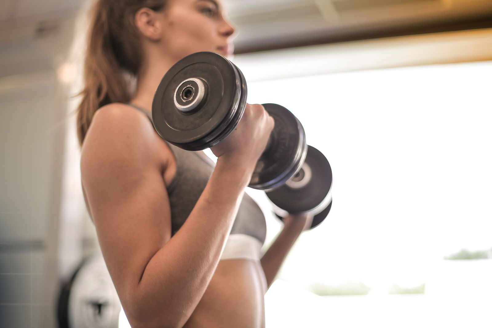 A woman in a grey sports top lifting two dumbbells in a bright gym