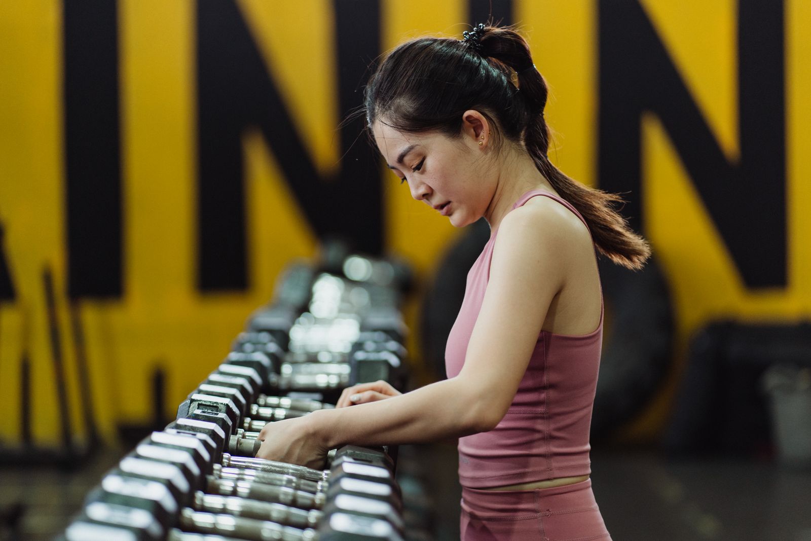 A woman in a pink training top choosing a dumbbell from a long rack in a gym