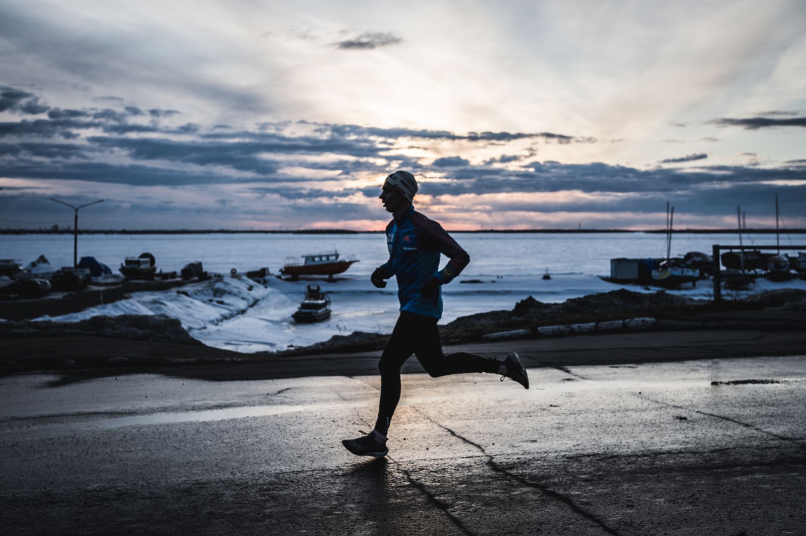 A runner in a hat and tights silhouetted on a waterfront road in winter, snow on the boats behind