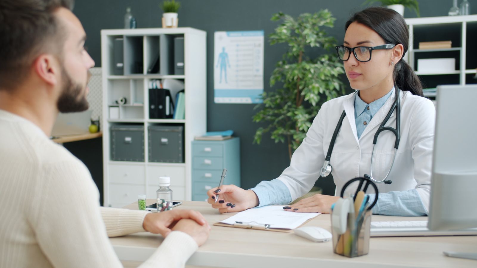 A surgeon in a white coat talking with a patient across a consulting desk, the patient seen from behind