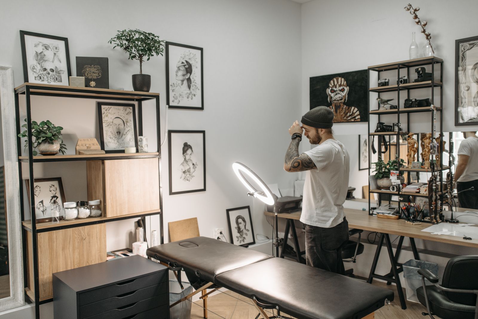 A tattoo studio room with a black client bed, framed drawings on the walls and shelves of supplies