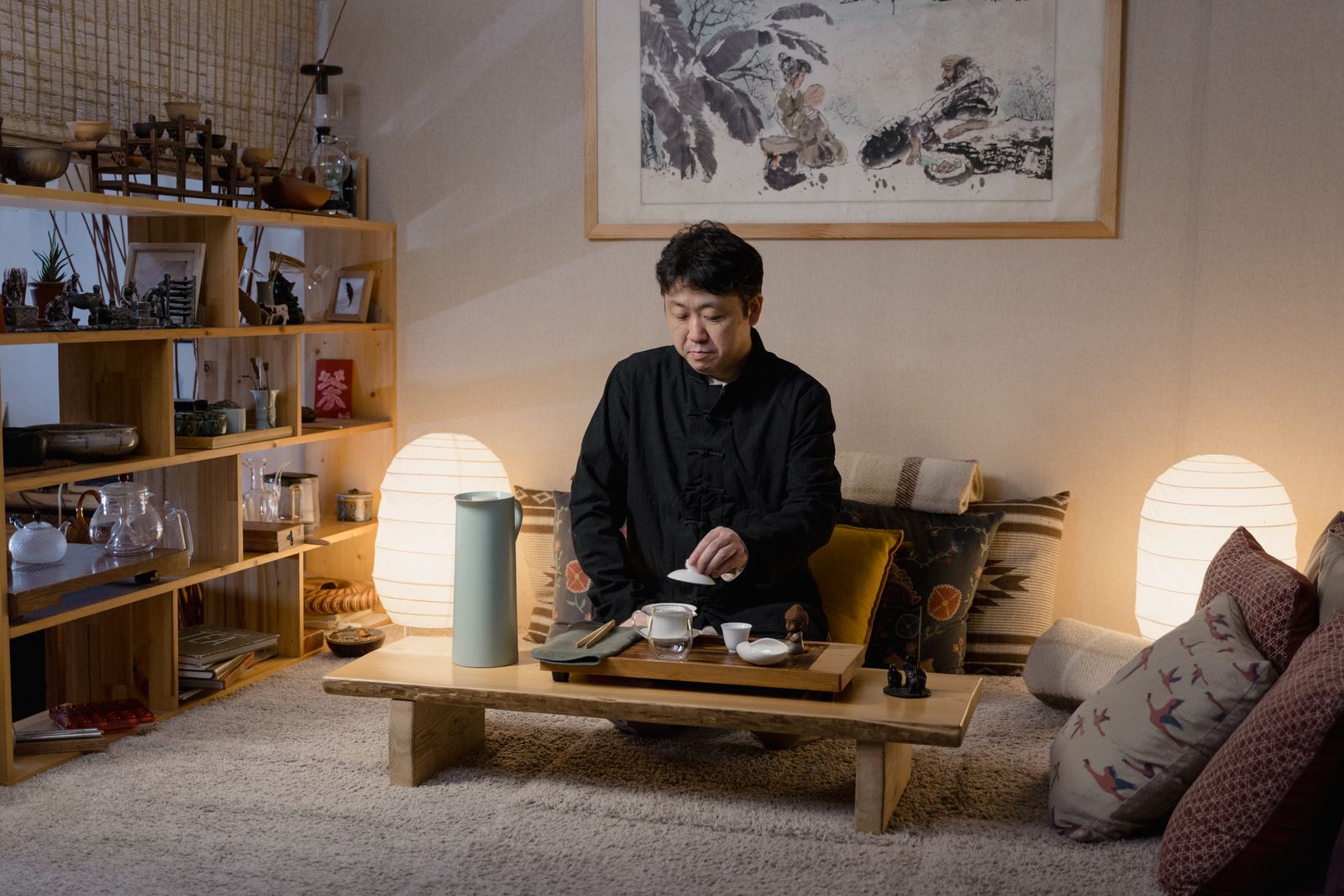 A tea host preparing tea at a low table in a quiet tea room with shelves of teaware