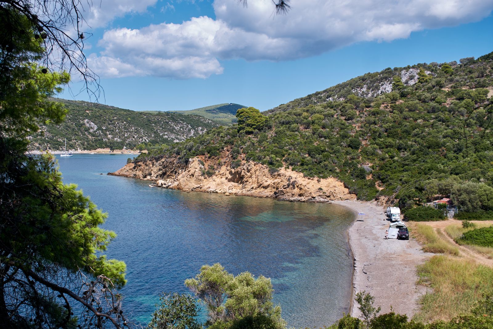 A quiet bay with green hills and a pebble beach seen through the trees
