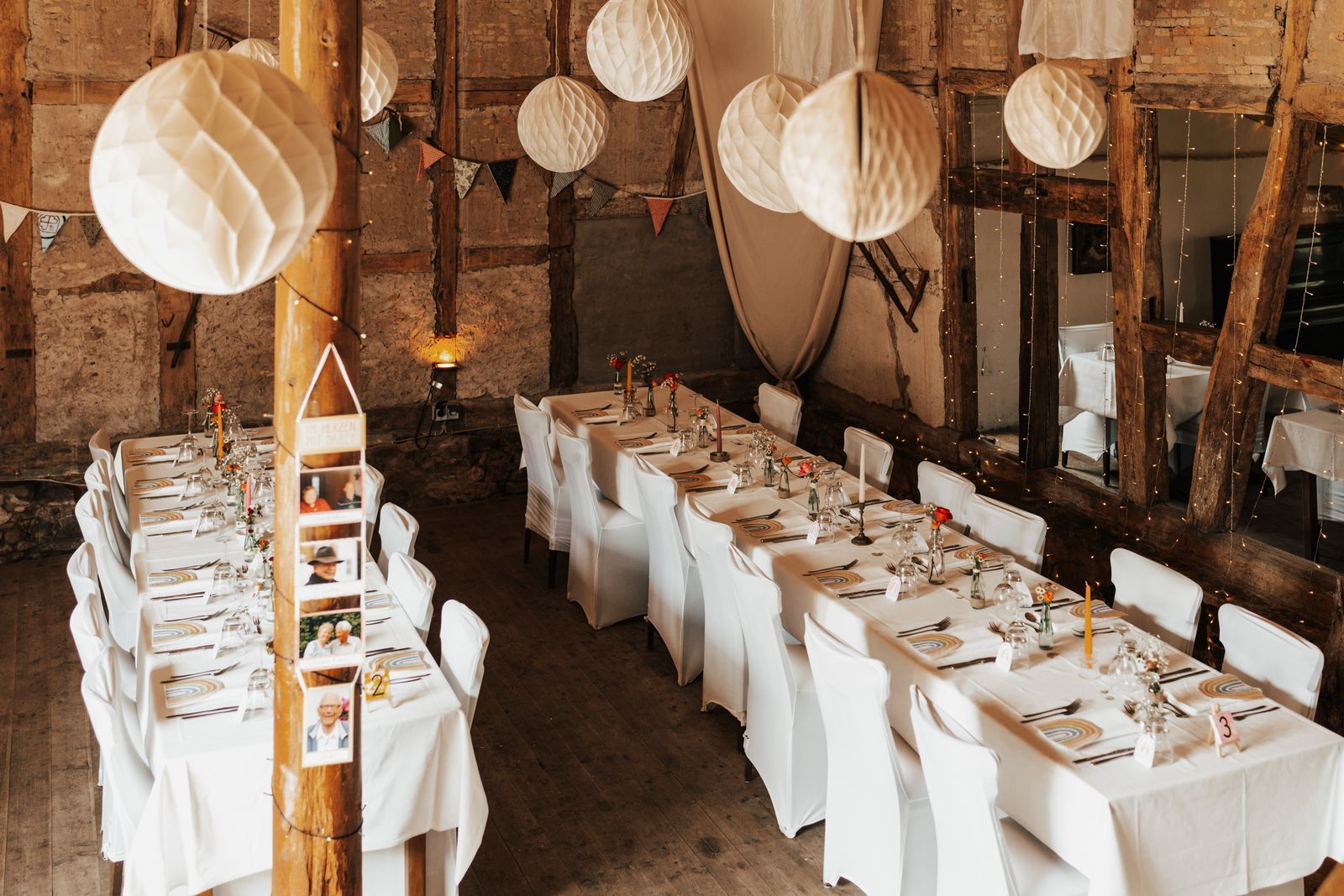 Long reception tables with white chairs under paper lanterns in a barn