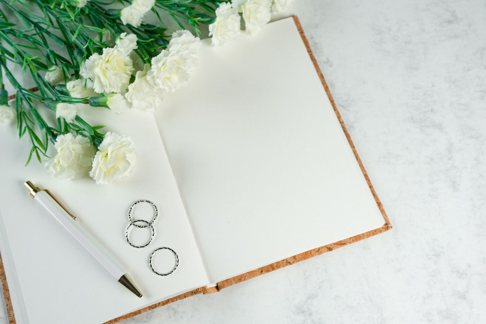 An open notebook with a pen, two rings and white flowers on a marble desk