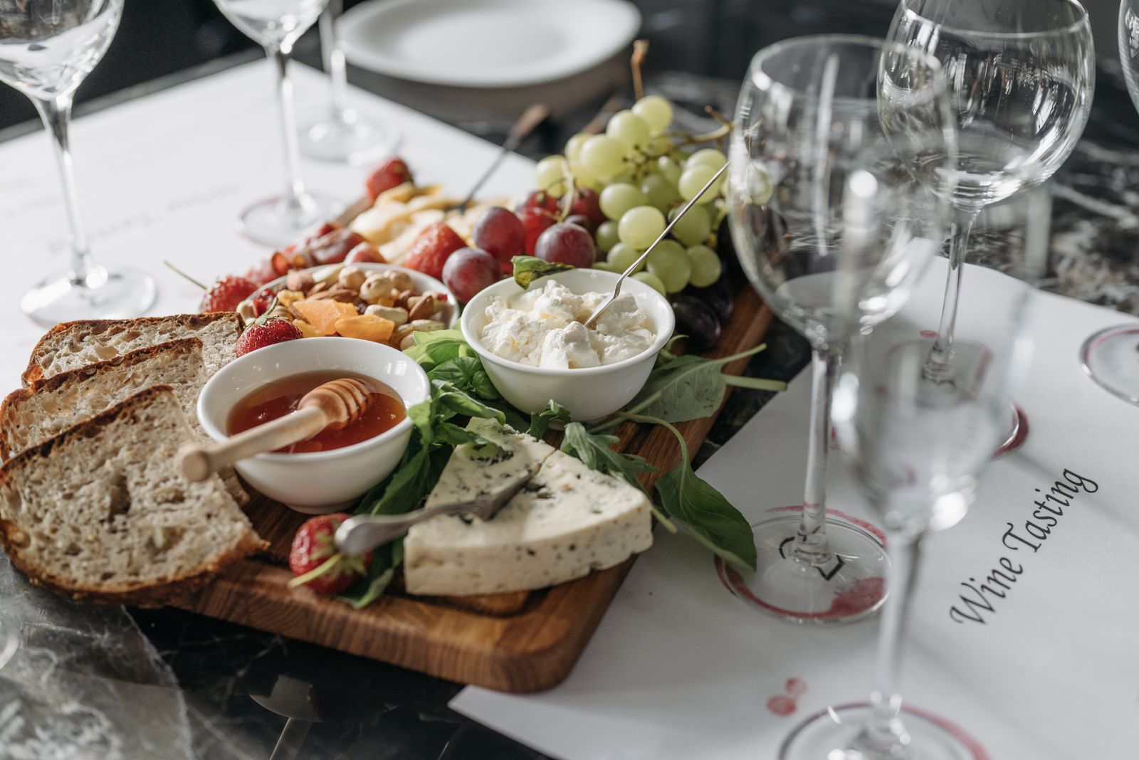 A board of cheeses, bread, fruit and honey beside empty wine glasses