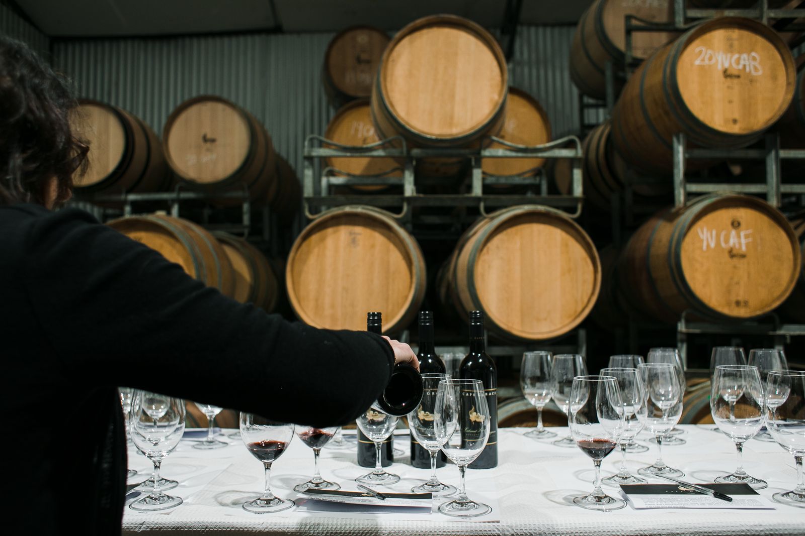 A host pours wine at a tasting table in front of oak barrels, seen from behind