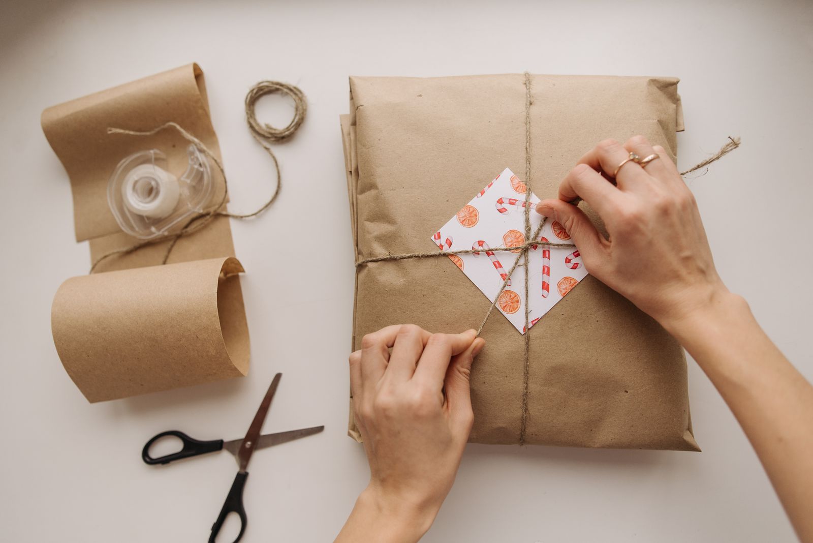 Hands tying string around a parcel wrapped in brown kraft paper, with scissors and a roll of paper